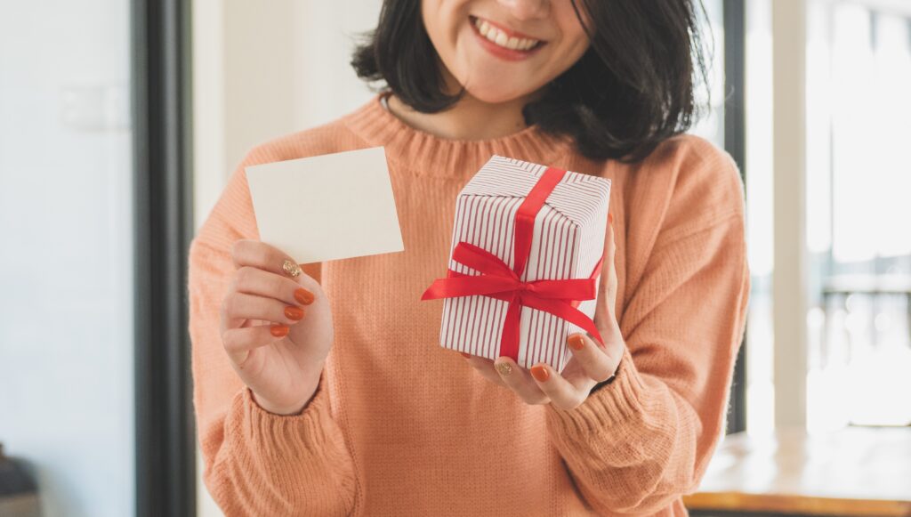 young asian woman in sweater holding striped gift box with red ribbon and blank wishing card